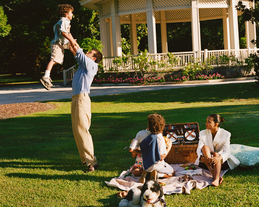 Family relaxing on a picnic blanket in a park with their dog, while the father playfully tosses his son into the air.