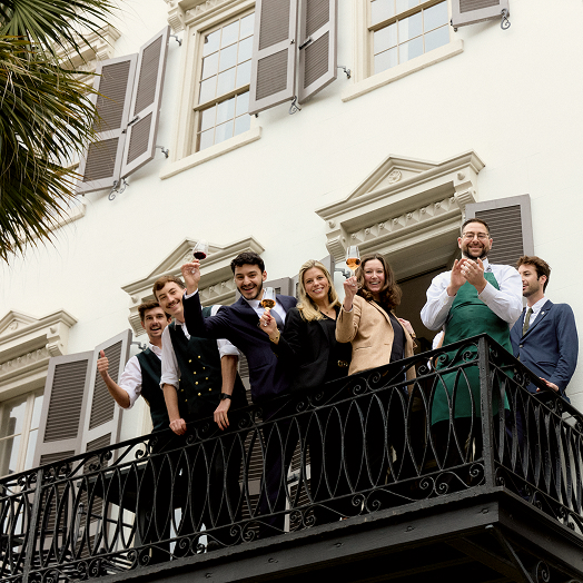 Hotel staff standing on a balcony smiling, holding drinks, and waving hello.